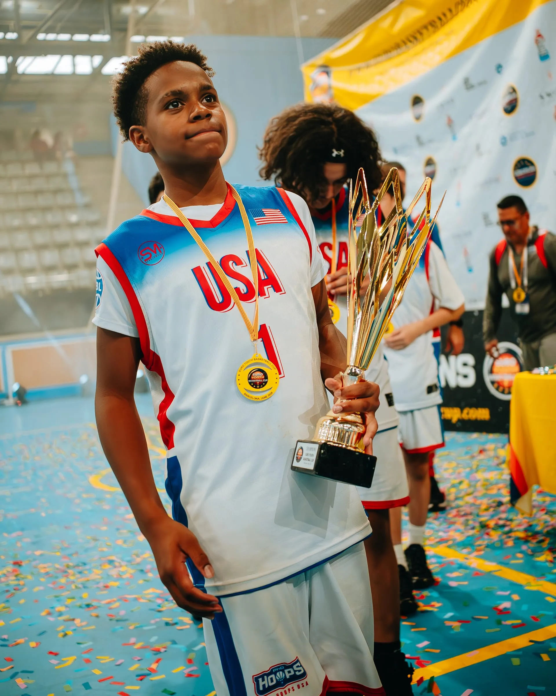 A young player wearing a gold medal and carrying a trophy across the confetti