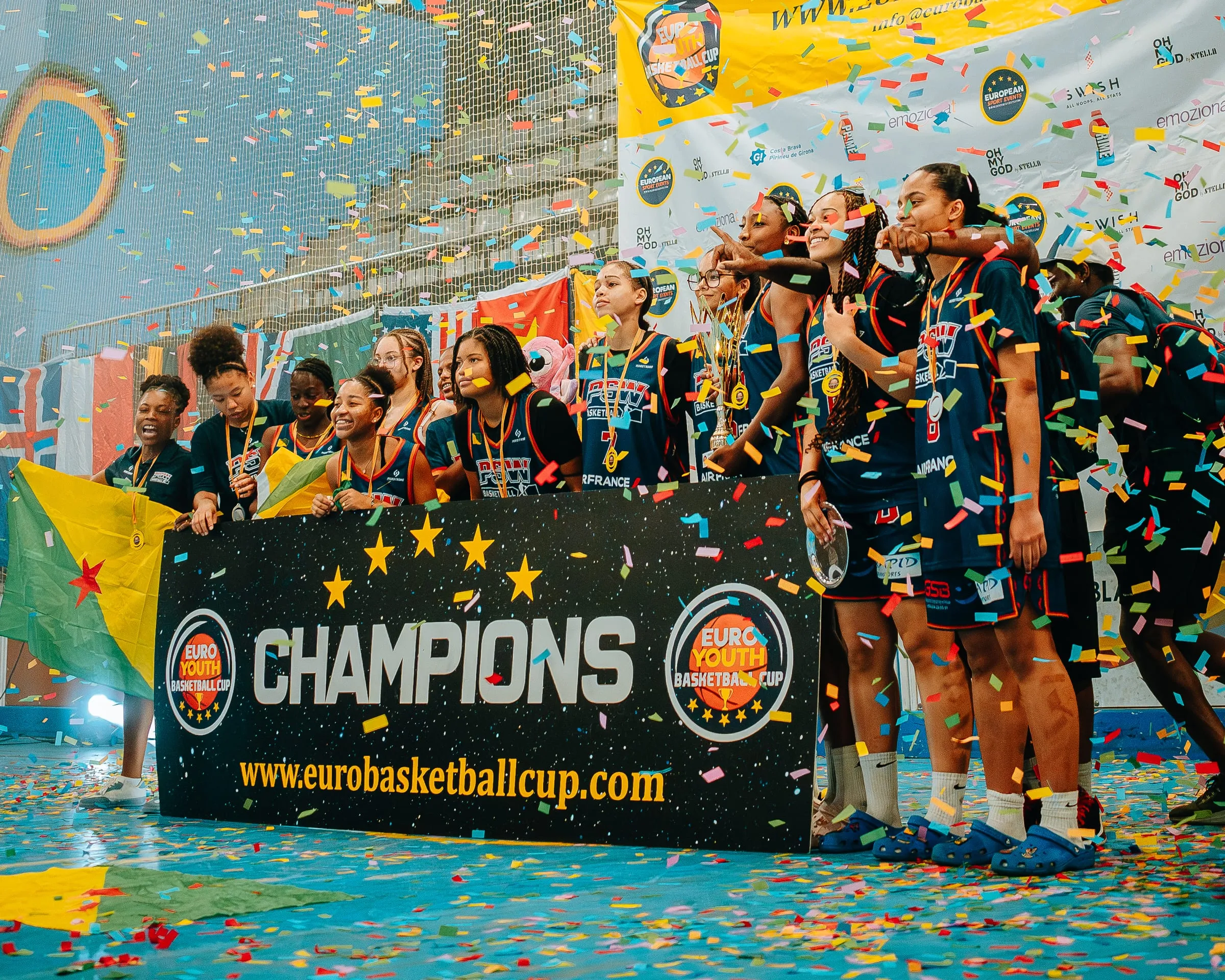 A team on the champions' podium under confetti, behind the Euro Youth Basketball Cup banner